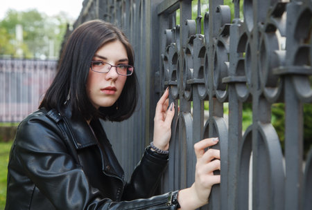 sad young girl in black jacket stands near metal fence on city street on summer day. closeup outdoorの写真素材
