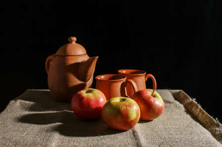 still life with a clay teapot, two cups and three red apples. indoor closeup on black backgroundの写真素材
