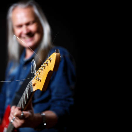 elderly man with long gray hair in blue denim shirt plays the electric guitar on black background