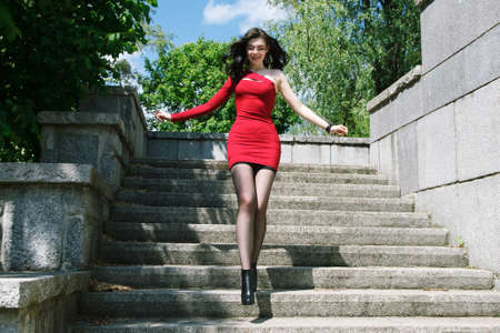 young brunette woman in a red dress goes down the stairs in city park on sunny summer dayの写真素材