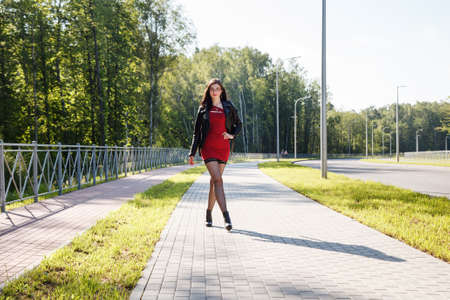 young brunette woman in red dress walking along the alley in the park on sunny summer dayの写真素材