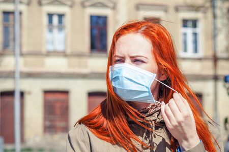Beautiful young European woman with ginger coloured hair on the street with a medical face mask on. Closeup of a young female in a respirator to protect against infection with coronavirus - Covid19の写真素材