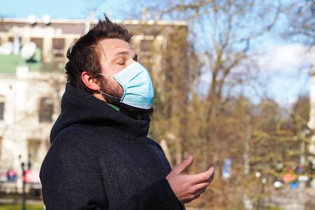 Handsome young European man coughing on the street with a medical face mask on. Closeup of a 35-year-old male in a respirator to protect against infection with influenza or coronavirus (Covid-19)の写真素材