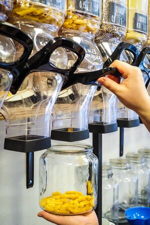 A woman holding jar with pasta with bulk dry food dispensers at zero waste shop in background,...