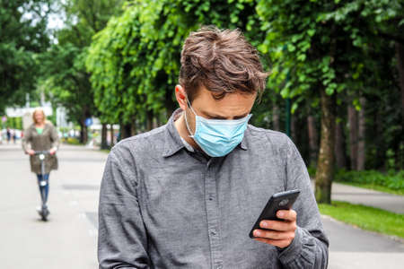 Handsome young European man with smartphone and a medical face mask on. Closeup of a 35-year-old male in a respirator to protect against infection with Covid-19の写真素材