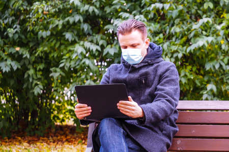 Handsome young European man in a park with laptop with a medical face mask on. Freelance working outside the office during an Covid-19 epidemic. Selective focusの写真素材