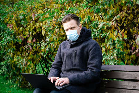 Handsome young European man in a park with laptop with a medical face mask on. Freelance working outside the office during an Covid-19 epidemic. Selective focusの写真素材