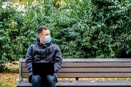 Handsome young European man in a park with laptop with a medical face mask on. Freelance working outside the office during an Covid-19 epidemic. Selective focusの写真素材