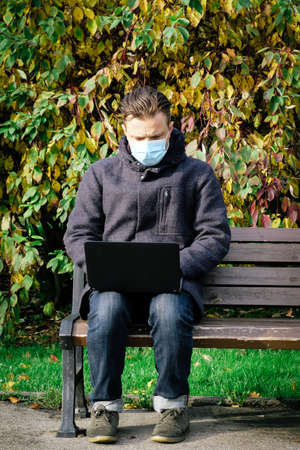 Handsome young European man in a park with laptop with a medical face mask on. Freelance working outside the office during an Covid-19 epidemic. Selective focusの写真素材