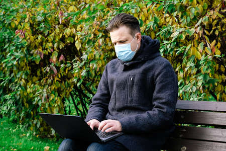 Handsome young European man in a park with laptop with a medical face mask on. Freelance working outside the office during an Covid-19 epidemic. Selective focusの写真素材