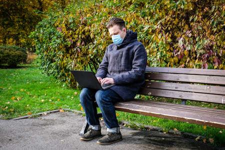 Handsome young European man in a park with laptop with a medical face mask on. Freelance working outside the office during an Covid-19 epidemic. Selective focusの写真素材