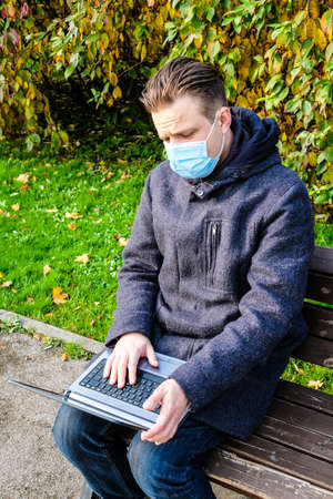 Handsome young European man in a park with laptop with a medical face mask on. Freelance working outside the office during an Covid-19 epidemic. Selective focusの写真素材