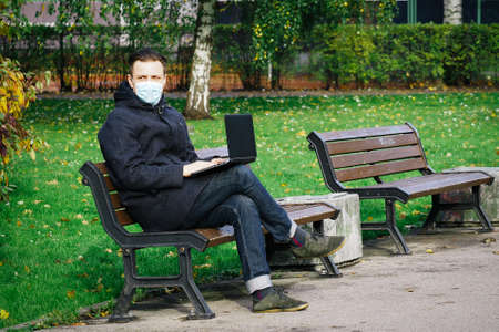 Handsome young European man in a park with laptop with a medical face mask on. Freelance working outside the office during an Covid-19 epidemic. Selective focusの写真素材