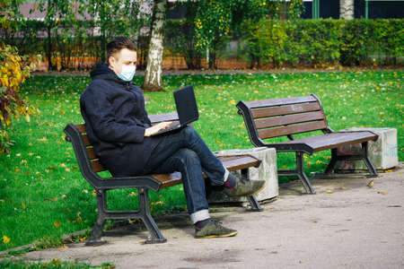Handsome young European man in a park with laptop with a medical face mask on. Freelance working outside the office during an Covid-19 epidemic. Selective focusの写真素材