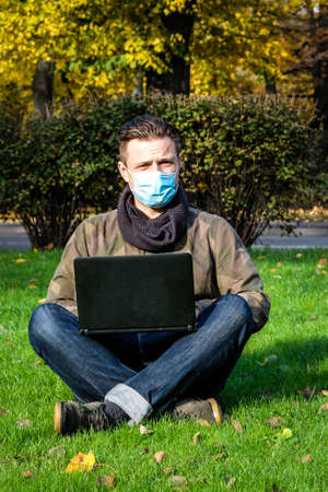 Handsome young European man in a park working at the computer lying on the grass in a protective medical mask on his face. Freelance, quarantine concept.の写真素材