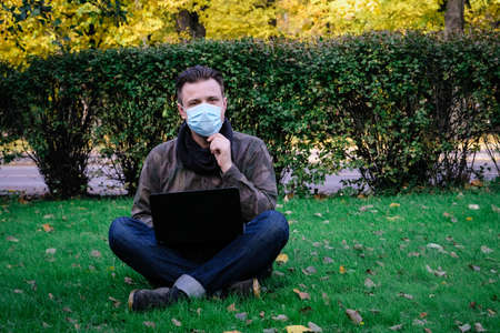 Handsome young European man in a park working at the computer lying on the grass in a protective medical mask on his face. Freelance, quarantine concept.の写真素材