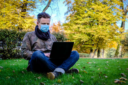 Handsome young European man in a park working at the computer lying on the grass in a protective medical mask on his face. Freelance, quarantine concept.の写真素材