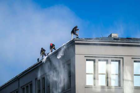 Riga, Latvia: February 12, 2021: Team of male workers clean roof of building from snow with shovels in securing belts. Selective focusのeditorial素材