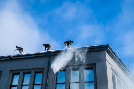 Riga, Latvia: February 12, 2021: Team of male workers clean roof of building from snow with shovels in securing belts. Selective focusのeditorial素材