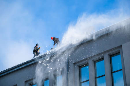 Riga, Latvia: February 12, 2021: Team of male workers clean roof of building from snow with shovels in securing belts. Selective focusのeditorial素材