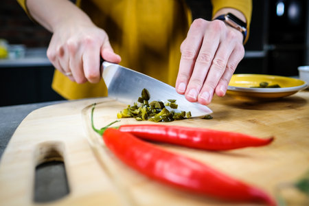 Woman cutting green spicy pepper jalapeno on wooden cutting board in the kitchen. Defocusedの写真素材