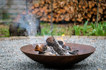 A close-up shot of burning firewood in a shallow, rusted steel fire pit placed on a gravel surface. Light smoke rises into the air while small flames dance around the blackened logs.の写真素材