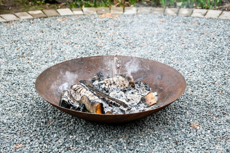 A close-up shot of burning firewood in a shallow, rusted steel fire pit placed on a gravel surface. Light smoke rises into the air while small flames dance around the blackened logs.の写真素材