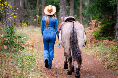 Young woman with long blonde hair wearing a cowboy hat, denim jacket, and fitted jeans walking with a saddled horse along a mossy forest trailの写真素材