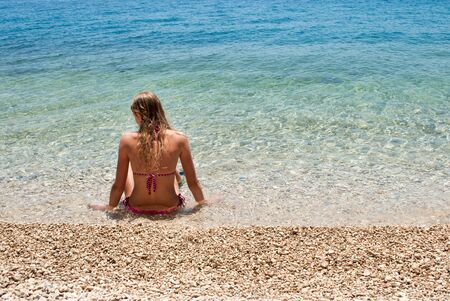 Young blond female sitting at the edge of a calm shallow blue sea, looking to the leftの写真素材
