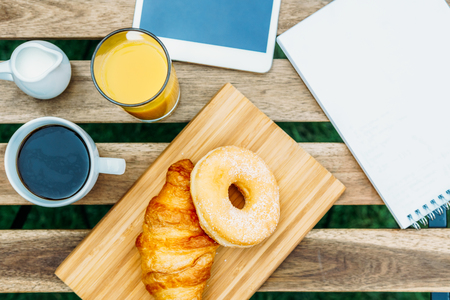 Morning Breakfast In Green Garden With French Croissant, Donuts, Coffee Cup, Orange Juice, Tablet and Notes Book On Wooden Tableの写真素材