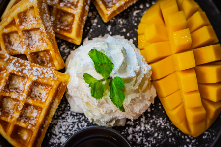 Whipped cream, mint leaf, mango and waffles sprinkled with powdered sugar served on a black plateの写真素材
