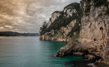 Cliffs of rock and vegetation in the sea, with cloudy storm sky and blue turquoise water in Cantabria Spainの写真素材