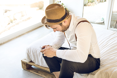 Guy modern dress looking at the phone sitting on the bed. Attractive and modern guy looking at the phoneの写真素材