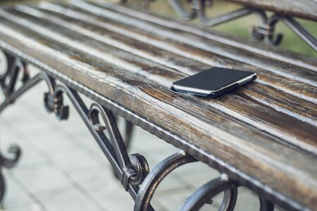 Left smartphone on a wooden bench in urban public park. Lost and forgotten personal thing outdoors in the cityの写真素材
