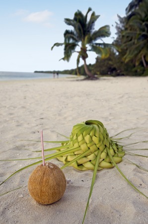Coconut with straw and sun hat knees out of palm leaves with couple of tourists in the background  on sandy sea shore of tropical island.の写真素材