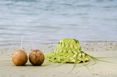 Two coconuts and sun hat knees out of palm leaves on sandy sea shore of tropical island.の写真素材