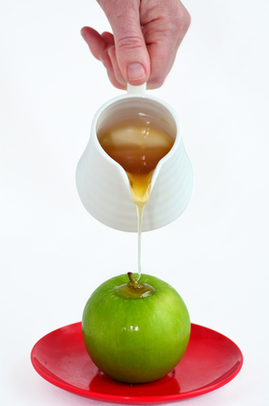 Jewish woman hand holds honey pot and pour fresh honey with on green apple during Rosh Hashanah Jewish holiday, on white background with copy spaceの写真素材
