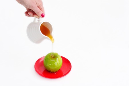 Jewish woman hand holds honey pot and pour fresh honey with on green apple during Rosh Hashanah Jewish holiday, on white background with copy spaceの写真素材