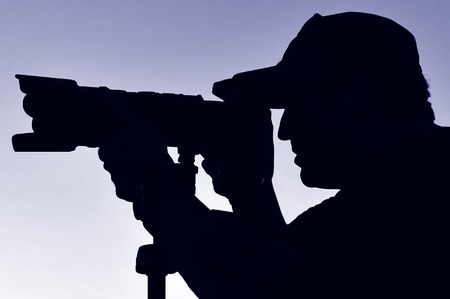 Silhouette of a photographer wearing a hat, camera and tripod with a blue sky background