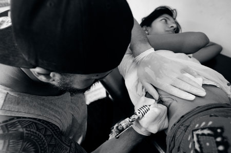 Pacific Island male tattoo artist draws a design on back of young woman client. real people 2 adult in tattoo  studio. (BW)のeditorial素材