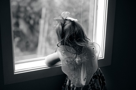 Sad little girl wearing angel fairy wings look outside from a home window.の写真素材