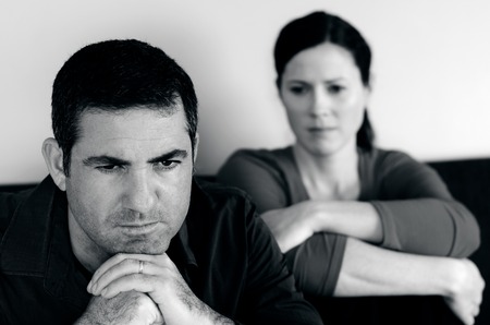 Portrait of unhappy young couple who have fallen out over a disagreement sitting on a sofa. Man in the front and the woman in the background.(BW)の写真素材