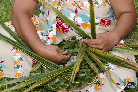 Close up of the hands of Polynesian Pacific Island Tahitian mature female weaving a hat knees out of palm leaves outside here home in Aitutaki lagoon Cook Islands.の写真素材