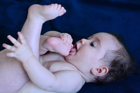 Baby lying on her back sucks toe on blue background.の写真素材