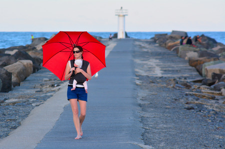 Young mother carry her infant baby and red umbrella in Seaway Gold Coast Australia.の写真素材