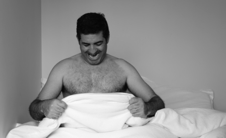 Happy man in his forties (40s) in bed looking down at his penis under white covers sheet in bedroom. Concept photo of male sexuality and man sex and relationship problem concept. (BW)の写真素材