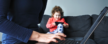 Mother works on laptop while her child plays on smartphone in the background. Woman works from home concept. copy spaceの写真素材