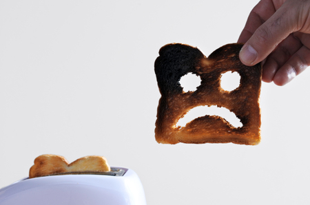 Hand holds one slices of burnt toast bread with sad face against a toaster. Sadness concept. copy spaceの写真素材