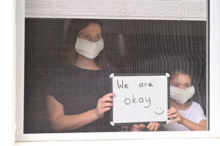 Mother and daughter (age 10)  in self isolation wearing a medical face mask looking outside home window holding a sign reading: We are okay to relatives.の写真素材