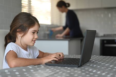 Young girl (age 10) using a laptop at home while her mother cleaning the kitchen in the background. Real people. Copy spaceの写真素材